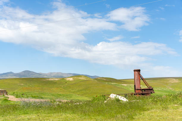 A rusty old structure sits in a grassy field. The sky is clear and blue, with a few clouds scattered throughout. The scene is peaceful and serene, with the old structure adding a sense of history