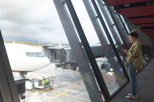 While waiting for her flight at the airport, a female tourist is using a smartphone and standing in front of the airport window
