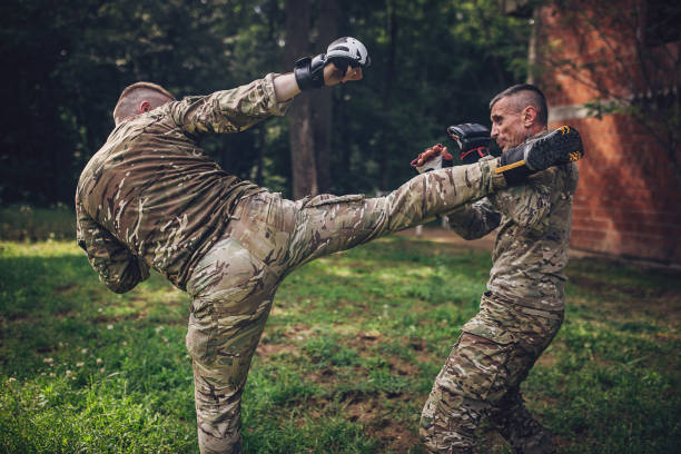Two military personnel practicing combat techniques outdoors, with one raising a leg to deliver a high kick while the other uses protective gear to block the kick.
