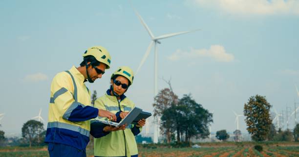 Renewable energy engineers are intently reviewing data on a laptop, contemplating the expansive turbine field before them. Green ecological power generation wind sustainable energy.