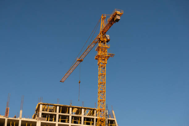 Towering yellow crane at a construction site against a clear blue sky, with a building under construction and visible scaffolding