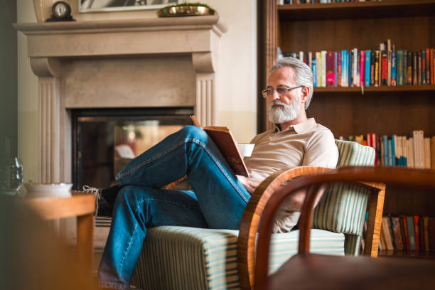 A smiling senior Caucasian man engages with a book while sipping coffee, relaxed in a striped armchair amidst a boutique hotel's cozy lounge, warm lighting enhancing the tranquil setting.