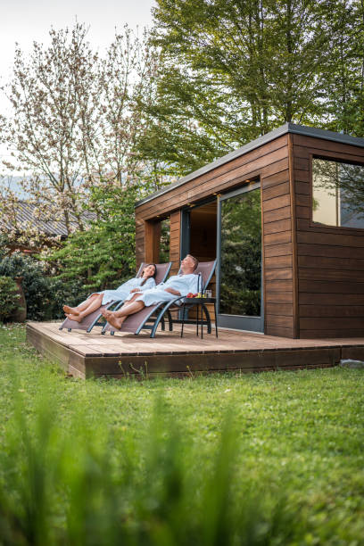 A couple lounges on deck chairs outside a wooden sauna cabin, wrapped in towels and basking in tranquility, surrounded by lush greenery.
