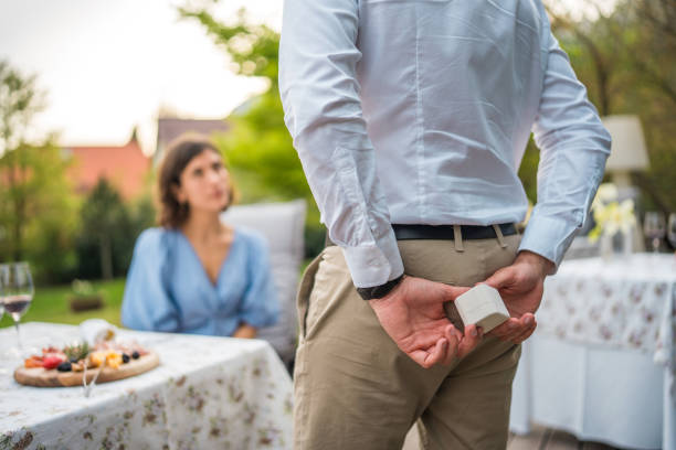 A mid adult Latin man discreetly holds an engagement ring behind his back, preparing to propose to his Caucasian partner. The couple sits at a decorated table, enjoying an intimate moment on a hotel terrace.