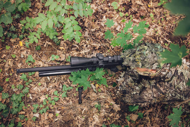 An overhead view showing a sniper in full camouflage gear aiming a precision rifle, blending seamlessly with the forest floor covered in leaves and greenery.