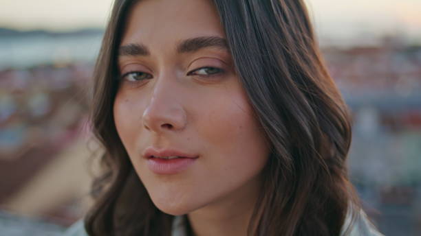 Romantic model posing cityscape rooftop in front sunset sky portrait. Relaxed serene brunette looking camera relaxing at balcony alone closeup. Tranquil woman resting terrace contemplating town scape