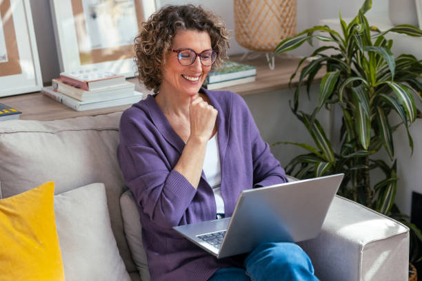 Kind mature woman using laptop while relaxing sitting on the couch at home