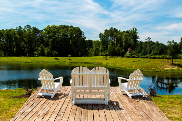 This image showcases a wooden dock patio set with white chairs arranged to face a serene pond. The setting is serene and inviting, with a clear blue sky and lush greenery providing a perfect backdrop for relaxation or outdoor activities.