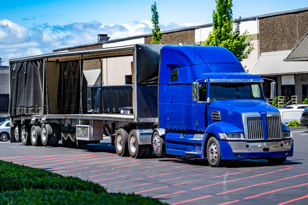 istock Blue big rig semi truck tractor with opened tented Conestoga flat bed semi trailer unloading delivered cargo standing on the industrial warehouse parking lot 2163722946