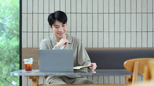 istock Pleasant asian man sitting at a table in a modern cafe working on a laptop and writing notes 2163729356