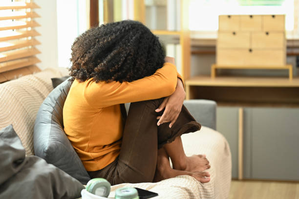 Sad upset young Afro woman hugging knees sitting on sofa, feeling lonely desperate. Mental health concept
