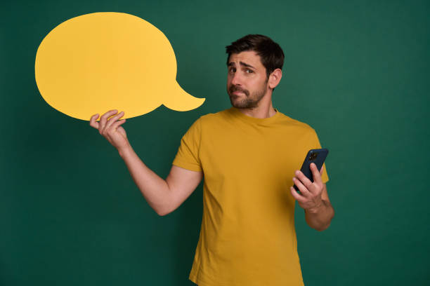 istock Young man holding speech bubble and smartphone looking confused 2163736850