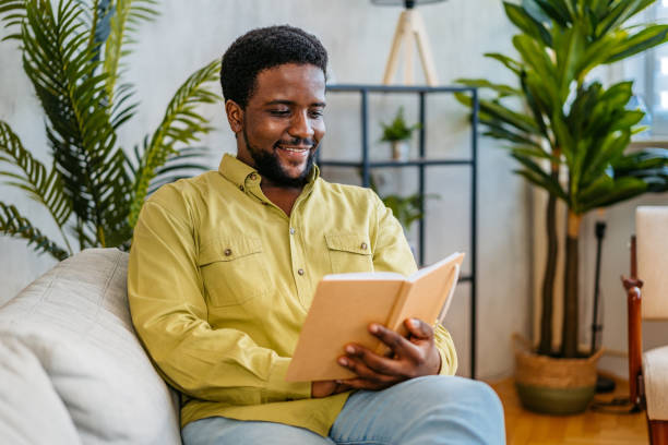 istock Young Man Reading A Book While Sitting On The Sofa At Home 2163737643
