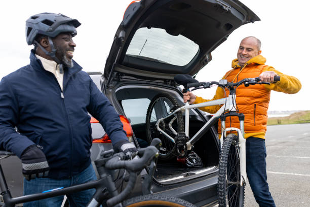 A medium-wide shot of two men unloading bikes from the boot of a car smiling at each other, they are both wearing jeans and winter coats. The man on the left is a polio survivor and lives with disabilities, they are in a car park at Whitley bay in the North East of England.Videos are available similar to this scenario.