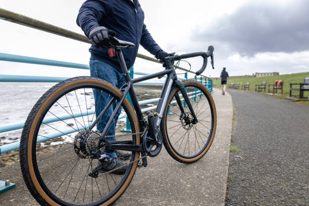 A wide shot of a man holding a road bike next to the beach. The man is wearing a blue jacket, he is in Whitley Bay in the North East of England.\n\nVideos are available similar to this scenario.