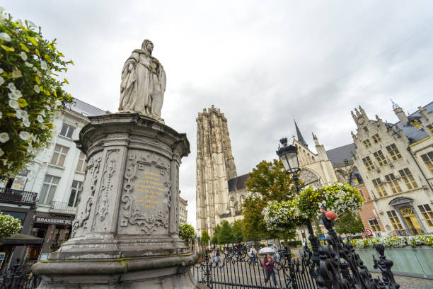 Discover the enchanting beauty of Mechelen, Belgium. This photo showcases the city's iconic Market Square, dominated by the towering Saint Rombouts Cathedral. The statue in the foreground and the floral decorations add a touch of elegance to this historic scene. The cloudy sky and the cobbled streets create a picturesque atmosphere, inviting you to explore this medieval gem.