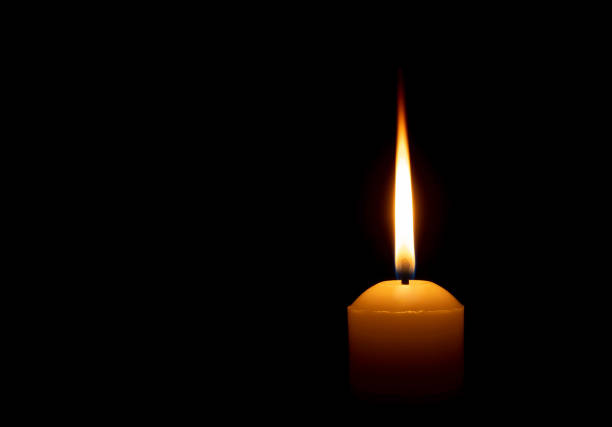 istock Single burning candle flame or light glowing on a big white candle on black or dark background on table in church for Christmas, funeral or memorial service with copy space. 2163783840