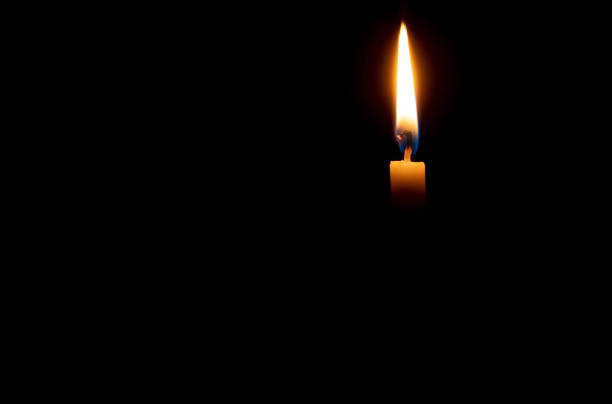 A single burning candle flame or light is glowing on an orange candle on black or dark background on table in church for Christmas, funeral or memorial service with copy space