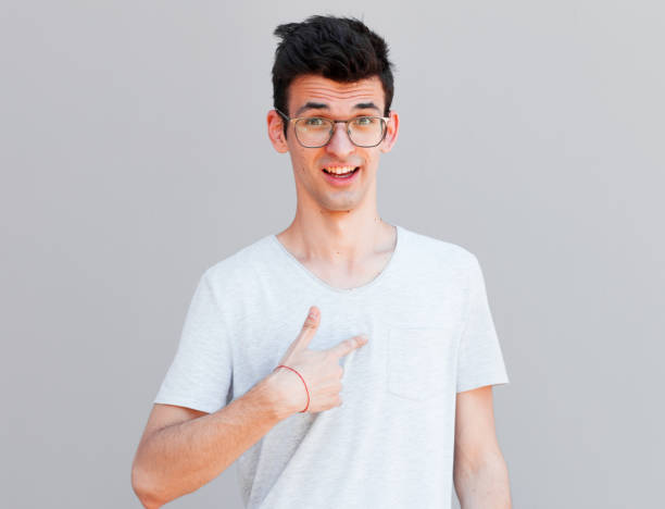 Studio shot of indignant man points at himself, feels embarrassed and displeased, points at himself, asks whether he is guilty, wears casual clothes, poses against grey studio wall. Who, me?