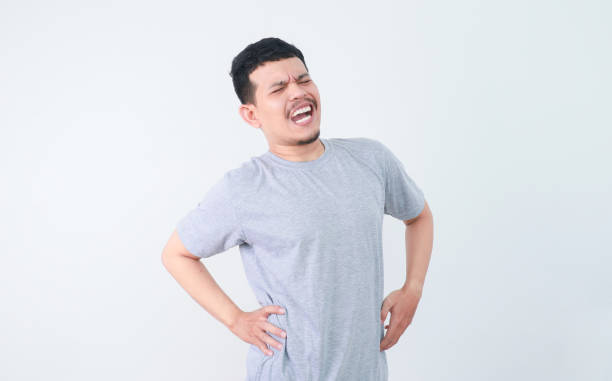 A man in a grey shirt bends over slightly and holds his lower back with a pained expression, indicating back pain or discomfort. health and welness concept.