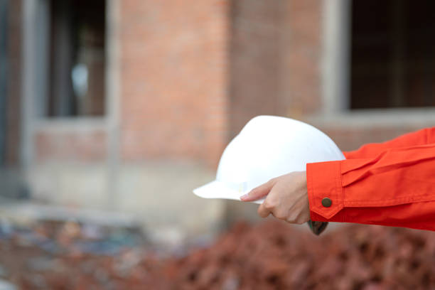 An engineer or foreman in orange coverall uniform is holding a white safety helmet or hardhat, standing in front of construction building site. Safety work practice in danger workplace concept.
