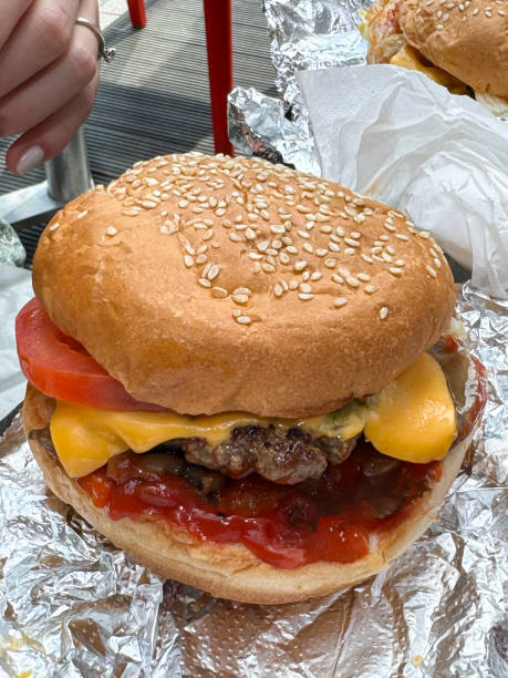 istock Close-up image gourmet cheeseburger on foil, sesame seed bun with beef hamburger patty, melted cheese slice, tomato slice, lettuce leaf, mushrooms, tomato ketchup sauce, al fresco fast food restaurant dining setting, woman's hand, focus on foreground 2163828097