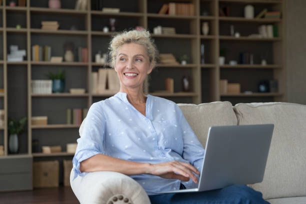 istock Smiling pensive mature woman using laptop on sofa at home 2163839883