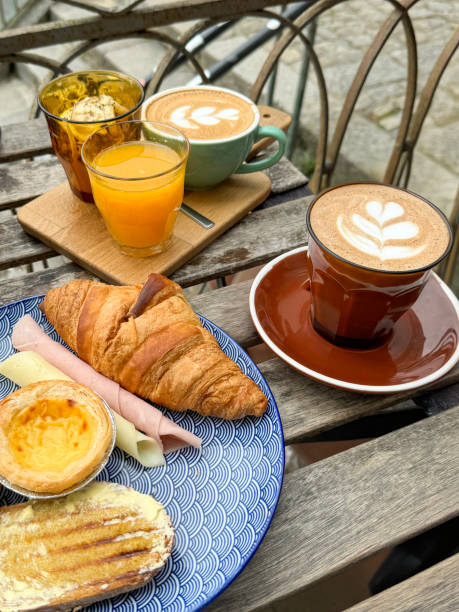 istock Close-up image continental breakfast on blue and white patterned plate, Pastel de nata (Portuguese egg custard tart), croissant, rolled ham, cheese slice, buttered toast, latte coffees, orange juice, al fresco dining scene, elevated view 2163842302