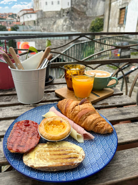 istock Close-up image continental breakfast on blue and white patterned plate, Pastel de nata (Portuguese egg custard tart), croissant, rolled ham, cheese slice, buttered toast with honey, latte coffees, orange juice, al fresco dining scene, focus on foreground 2163842447