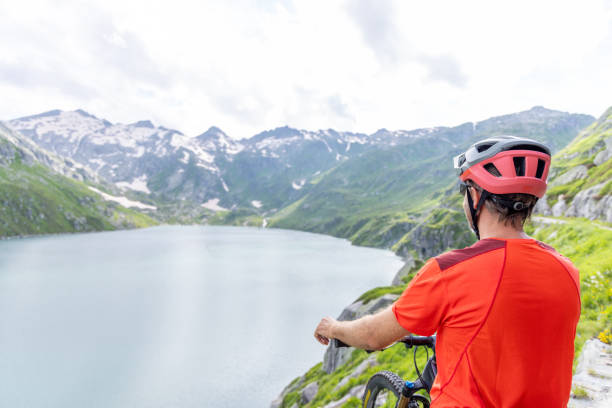 A man in biking gear stands with his mountain bike on a scenic path, taking in the breathtaking view of a tranquil mountain lake surrounded by snow-capped peaks and lush greenery.