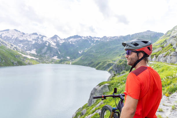 A man in biking gear stands with his mountain bike on a scenic path, taking in the breathtaking view of a tranquil mountain lake surrounded by snow-capped peaks and lush greenery.