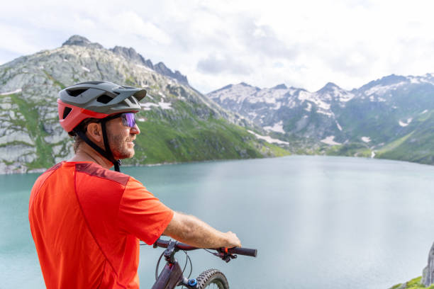 A man in biking gear pauses to admire the breathtaking view of a tranquil mountain lake, surrounded by snow-capped peaks and lush greenery.