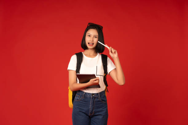 istock Asian short-haired female college student studying at university, taking photos in studio on red background, using laptop and pen to prepare for final exam, carrying bag, wearing glasses 2163899767
