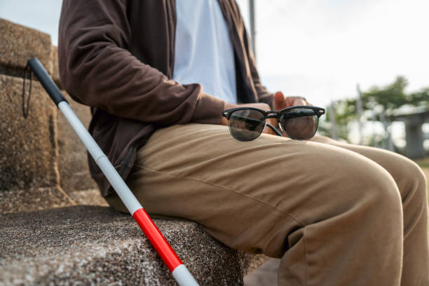 Young asian blind man sitting outside holding walking stick Visually impaired man difficult to traveling on the road wearing sun glasses Cross the road cross the footbridge stock photo