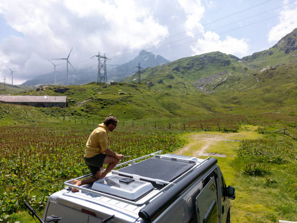 A man adjusts solar panels on the roof of his camper van, set in a lush green mountainous area. Wind turbines and power lines in the background highlight the integration of renewable energy sources.
