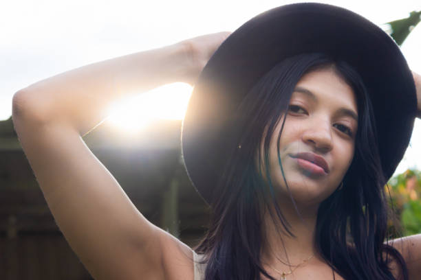 Beautiful young latina woman wearing black hat and looking at camera with cloudy sky in background