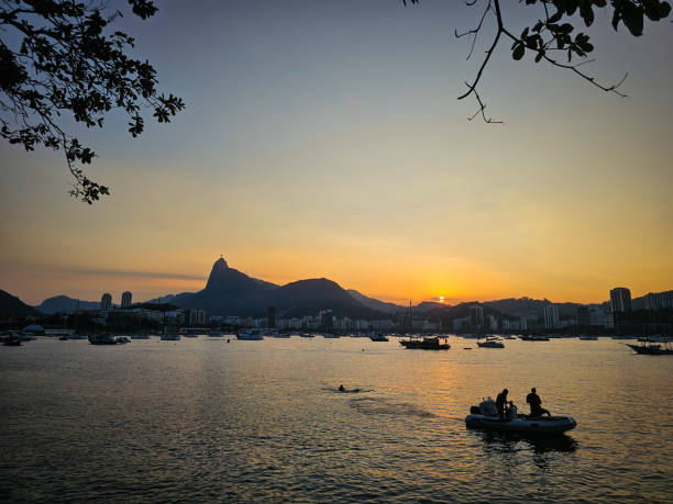 A breathtaking sunset view captured from Mureta da Urca in Rio de Janeiro, Brazil. The iconic silhouette of Corcovado Mountain is prominently visible against the backdrop of a golden sky. The calm waters of Guanabara Bay are dotted with boats and a small inflatable raft carrying two people, creating a tranquil and picturesque scene. The branches of a tree frame the image.