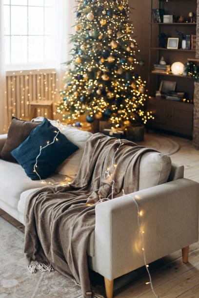 istock Sofa with pillows and a plaid on a blurred background with a Christmas tree decorated with garland lights in the interior of a cozy living room with large windows and brown walls. Warm Christmas Eve 2164013923