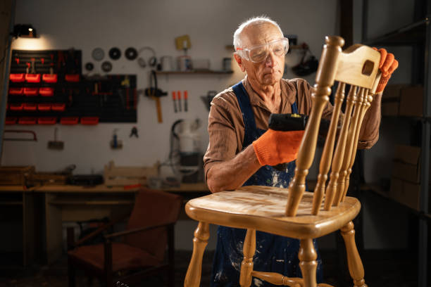 istock An elderly man in a workshop uses a drill with a special attachment to remove old varnish from a wooden chair. He is wearing protective glasses and orange gloves. 2164017674
