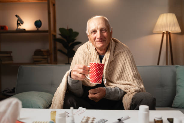 istock Hot drinking. Shivering elderly man catching cold suffering influenza holding cup healing grippe sitting wrapped blanket on couch and smiling on camera 2164017747