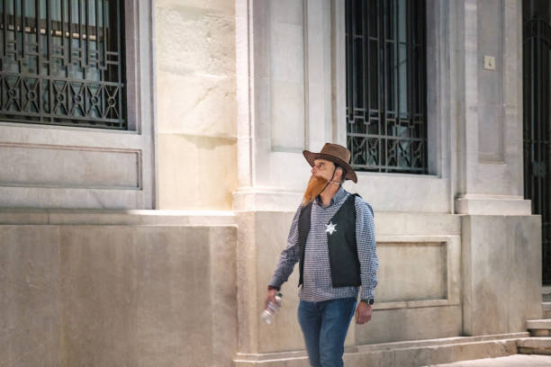 Man wearing a wide-brimmed hat, a mask or facial covering that features a beard design, and a star badge on their chest. denim jacket, a grey hoodie, and blue jeans, classical architectural style, with windows protected by bars and detailed stonework Malaga Spain 02/11/2024