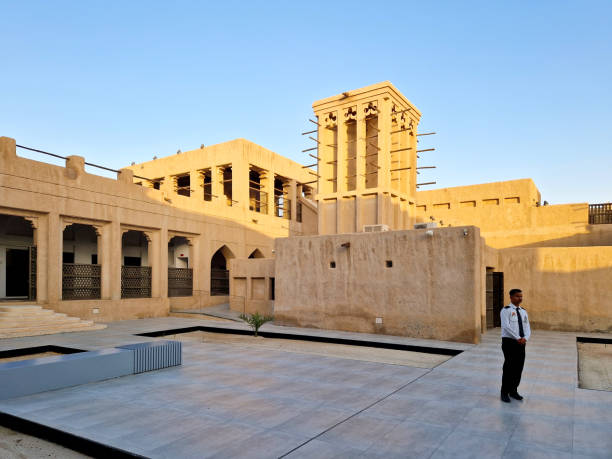The image shows a traditional courtyard in Deira, Dubai, featuring earthen buildings with a central wind tower (Barjeel). A security guard stands in the modernized courtyard, blending traditional architecture with contemporary elements under a clear blue sky.