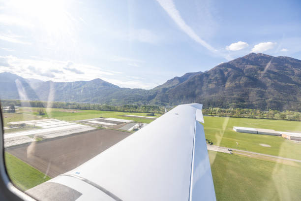 A light aircraft is seen on an airfield, with its wing in the foreground. The scene captures a sunny day with mountains and a few clouds in the background.