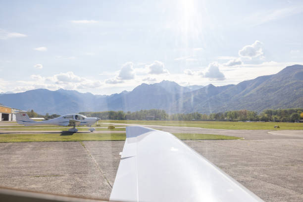 A light aircraft is seen on an airfield, with its wing in the foreground. The scene captures a sunny day with mountains and a few clouds in the background.