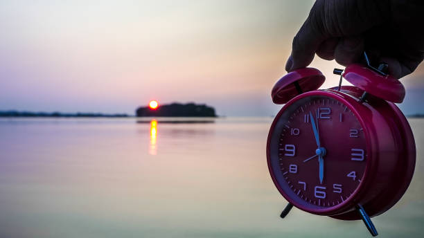 istock Sunset Over Water With Alarm Clock Held by a Hand at Dusk 2164076299