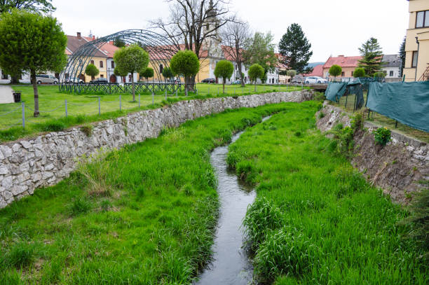 A small creek flows through a town, bordered by grassy banks and stone walls. The scene includes neatly trimmed trees and quaint buildings, adding charm to this peaceful and picturesque setting.