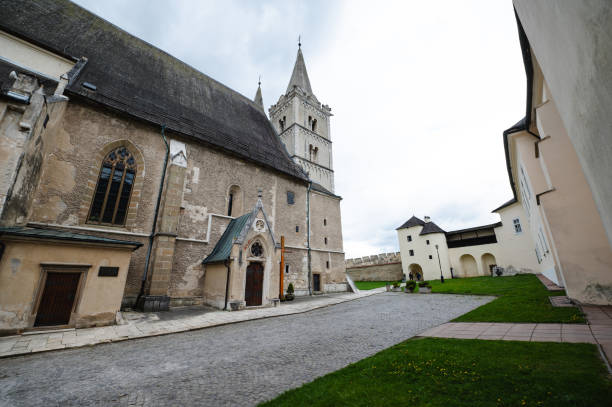 A historic church featuring twin spires and Gothic architectural elements, set against a cloudy sky. The church is surrounded by green lawns and adjacent buildings, creating a serene and picturesque scene.