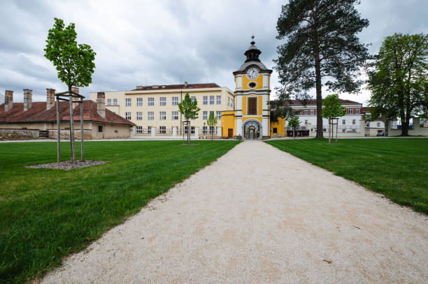 A charming yellow clock tower stands beside a gated entrance, leading to a scenic courtyard. The scene includes cobblestone streets, historic buildings, and a backdrop of green hills under a cloudy sky.