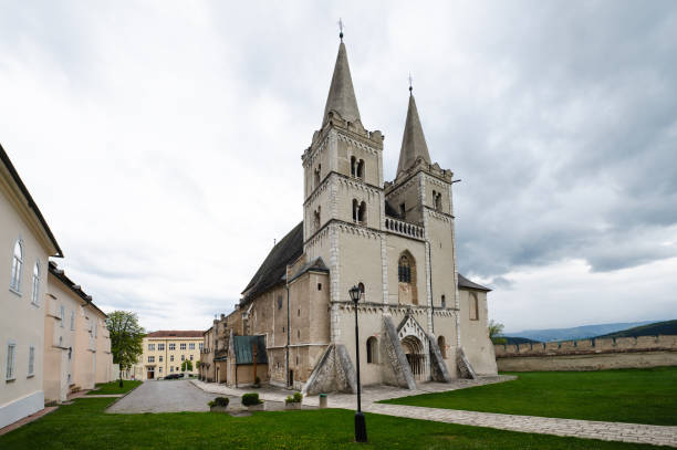 A historic church featuring twin spires and Gothic architectural elements, set against a cloudy sky. The church is surrounded by green lawns and adjacent buildings, creating a serene and picturesque scene.