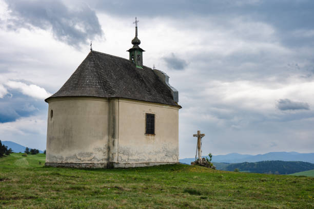 A historic chapel with a prominent crucifix stands atop a hill, framed by a dramatic cloudy sky. The building's weathered facade and simple architecture add to its serene and solemn atmosphere.